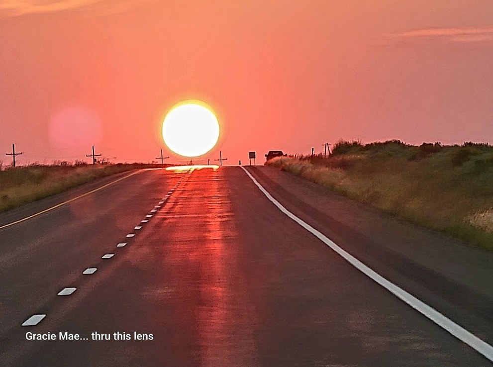 A glowing sun setting at the end of a long, empty highway under a pink sky